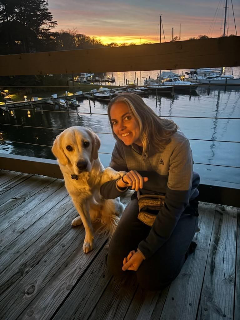 Woman kneels beside a yellow Labrador on a dock at sunset, with boats and water behind them.