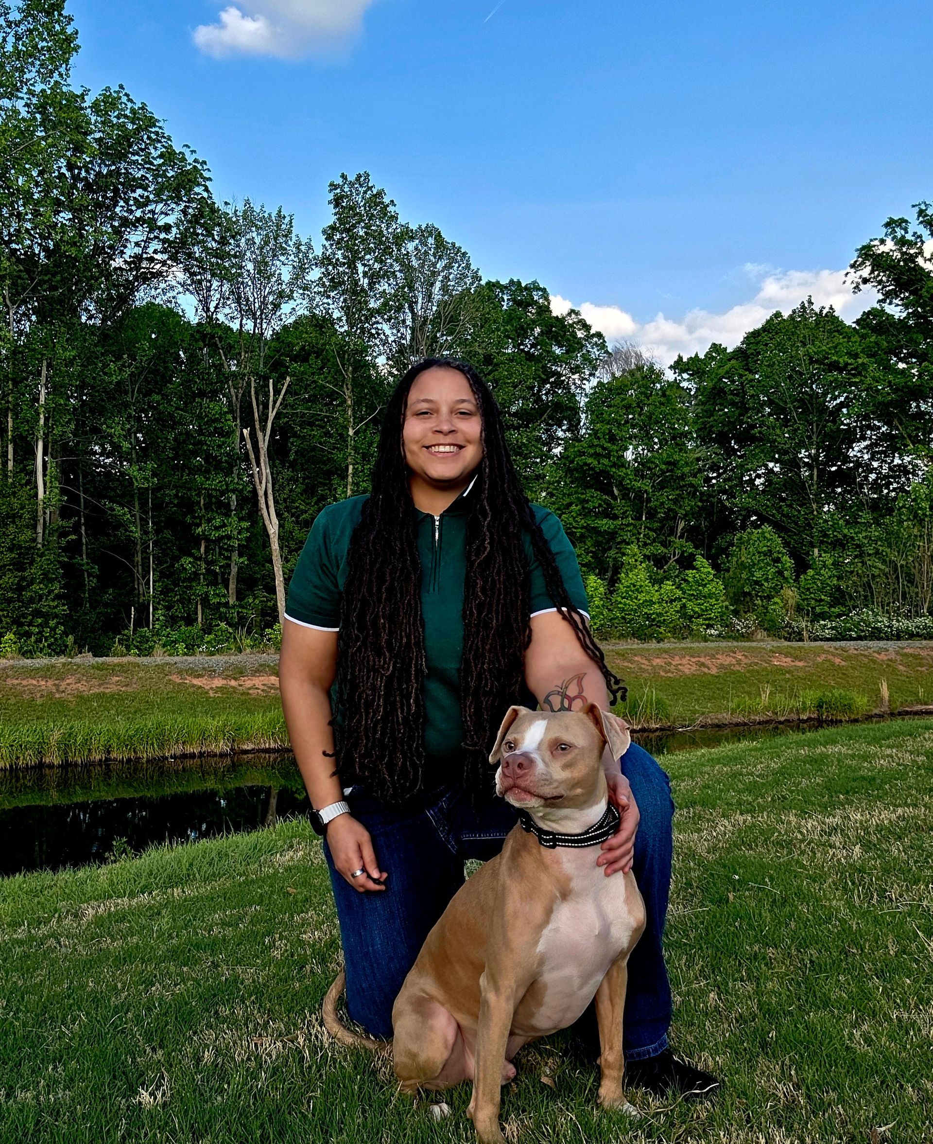 Woman standing beside a tan dog in a green park with trees and water in the background