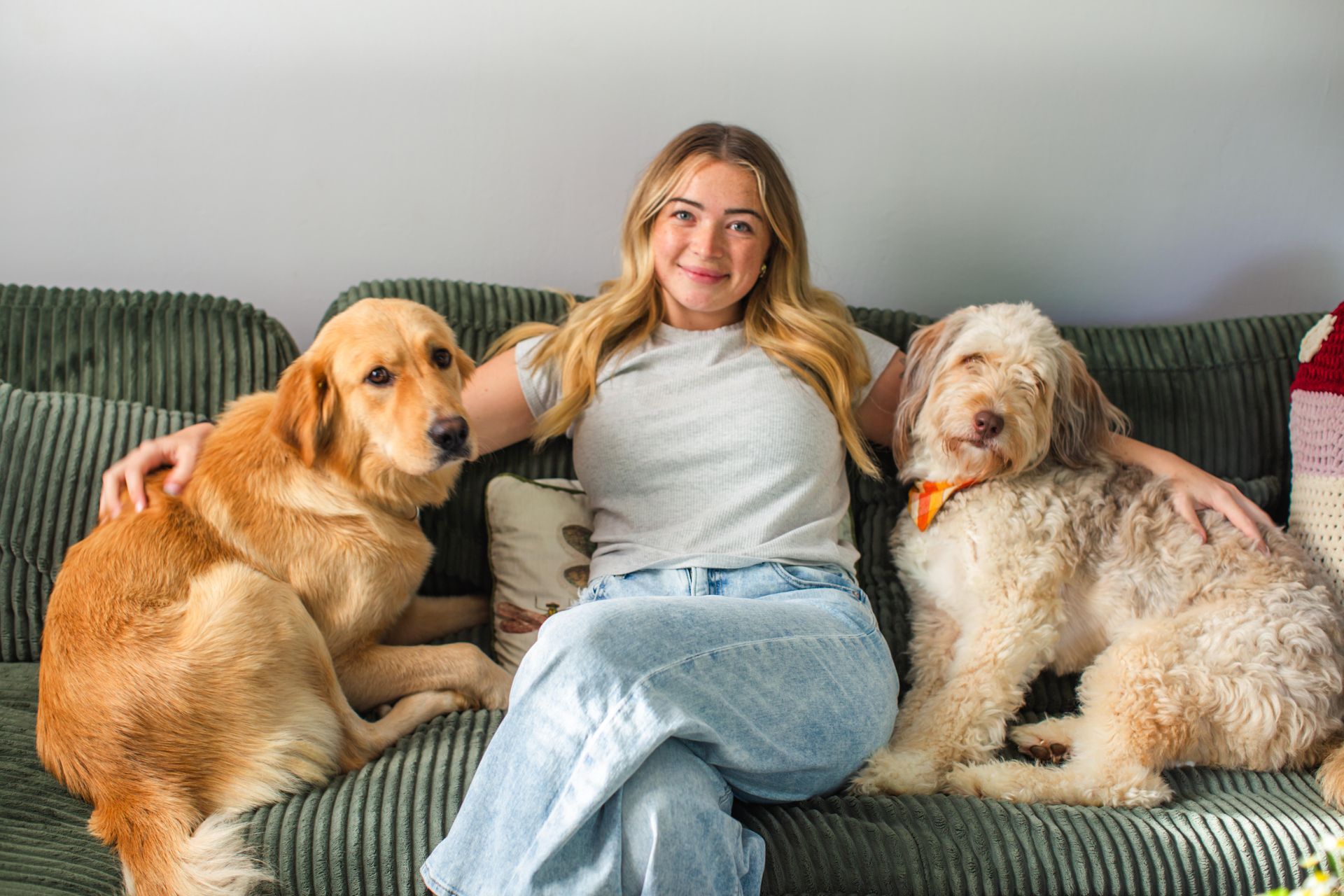 Woman sitting on a green couch with two dogs, smiling in a bright living room.