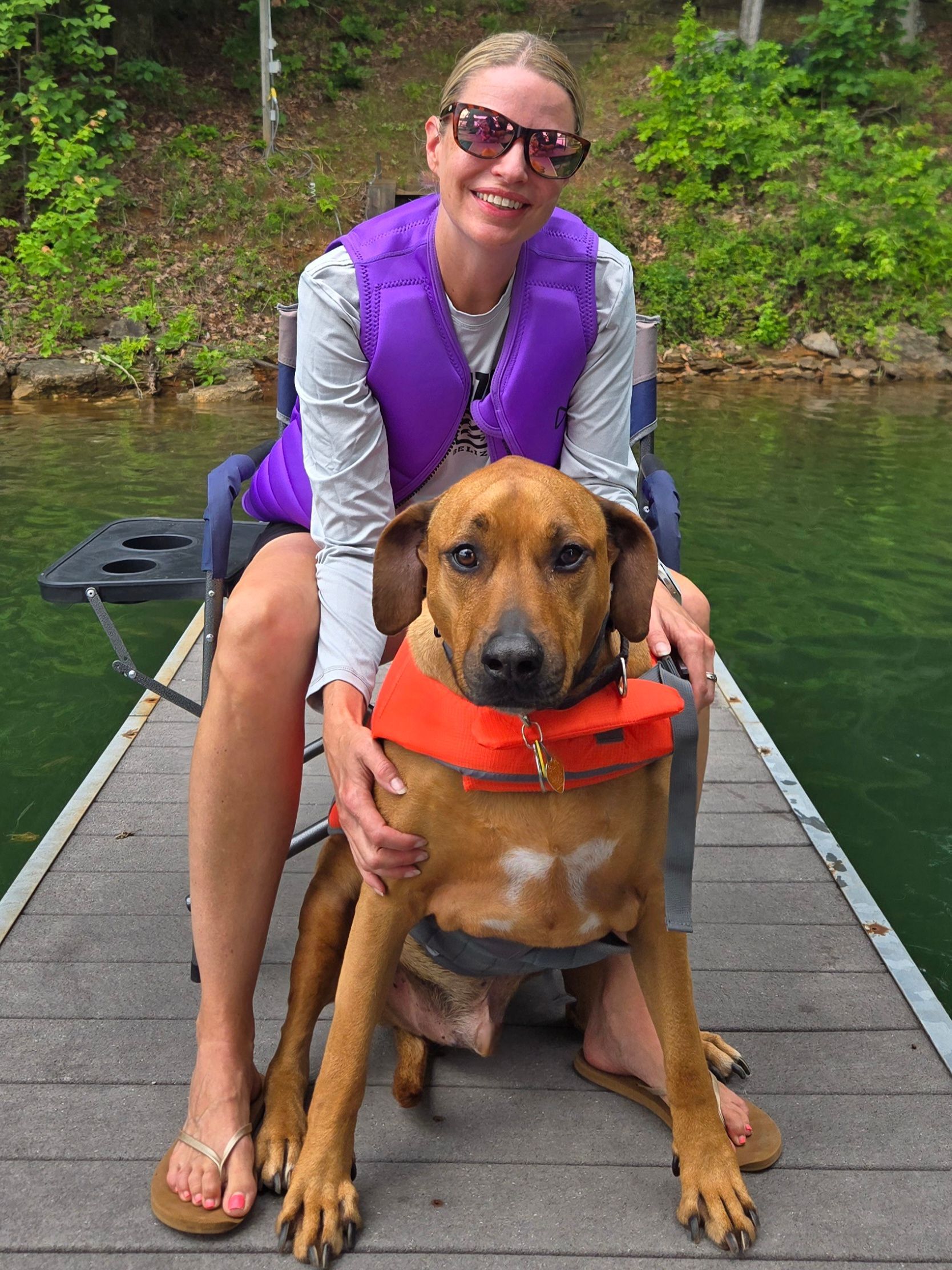Woman in a purple vest sitting on a dock with a dog wearing an orange life jacket by the water