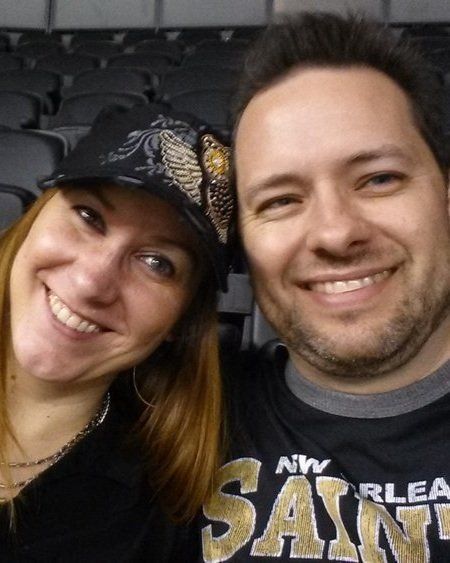 Two smiling people posing for a selfie indoors, one wearing a black cap and the other a dark T-shirt.