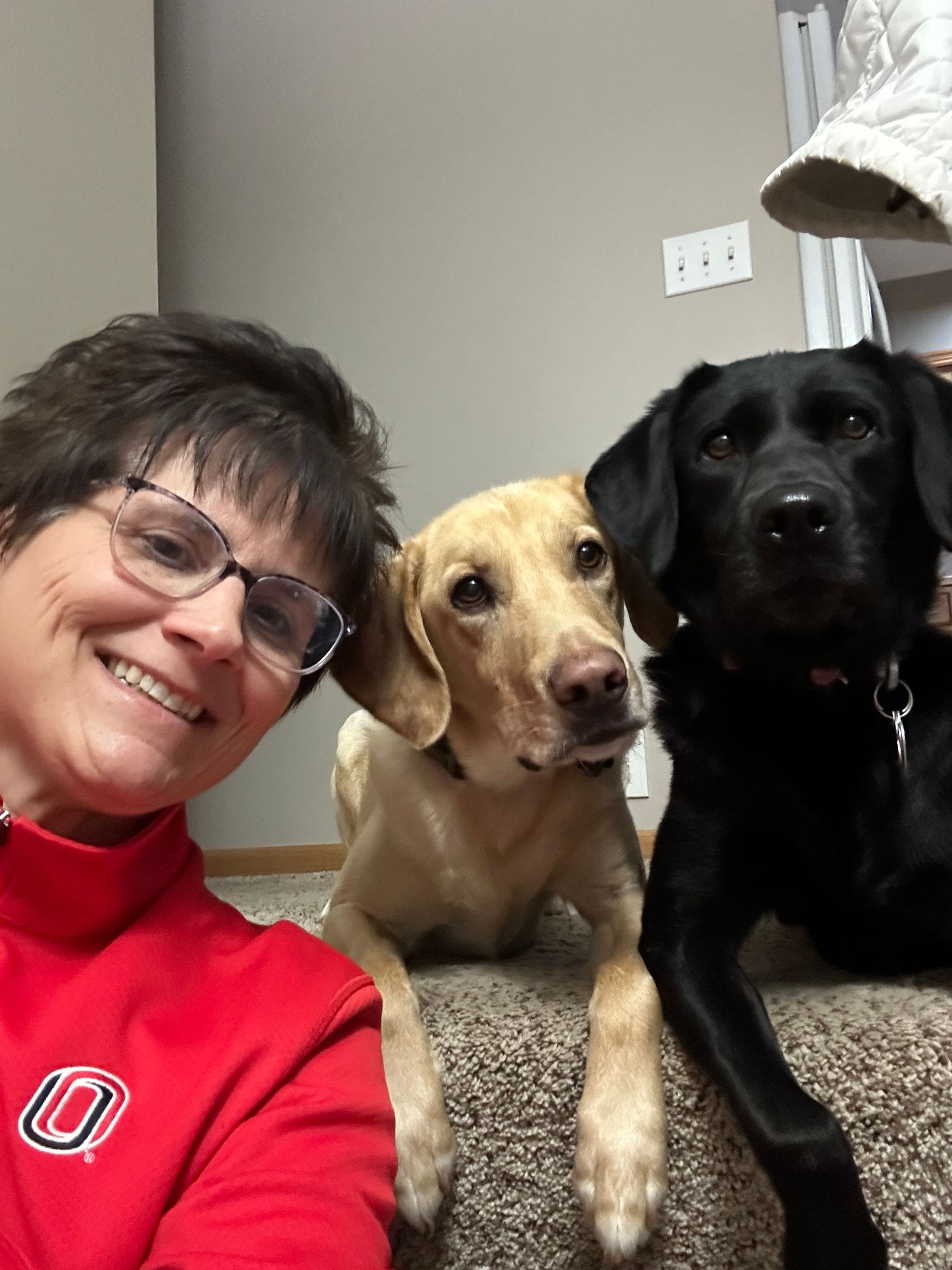 Woman in red shirt posing with two dogs, tan and black, indoors.