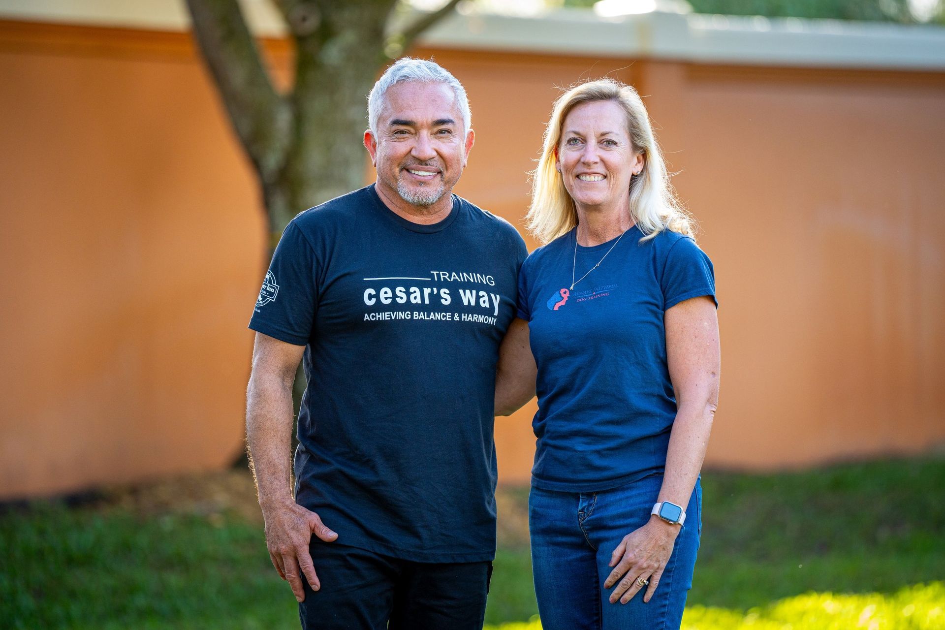 Two people standing outdoors, smiling, wearing navy shirts in front of a grassy wall.