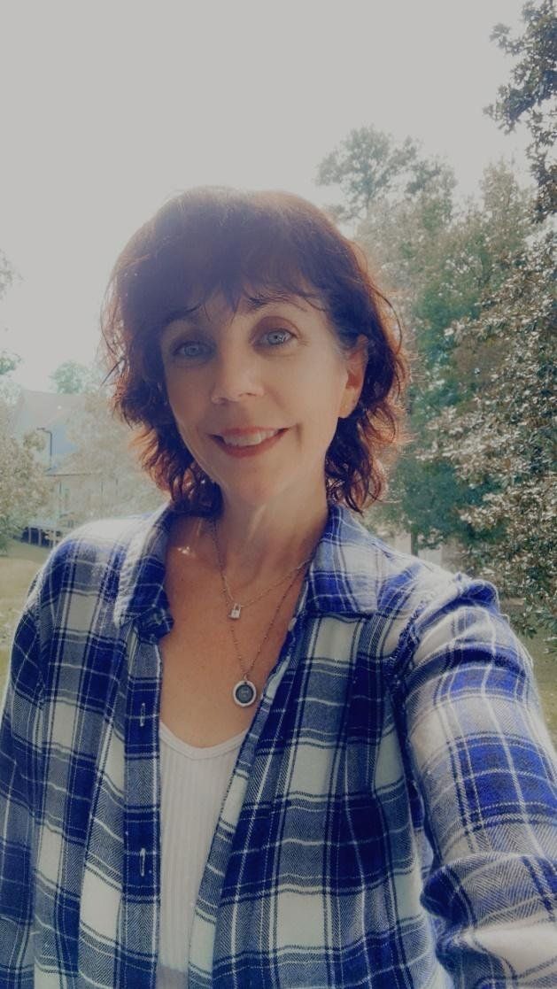 Smiling person in a blue plaid shirt standing outdoors with trees in the background