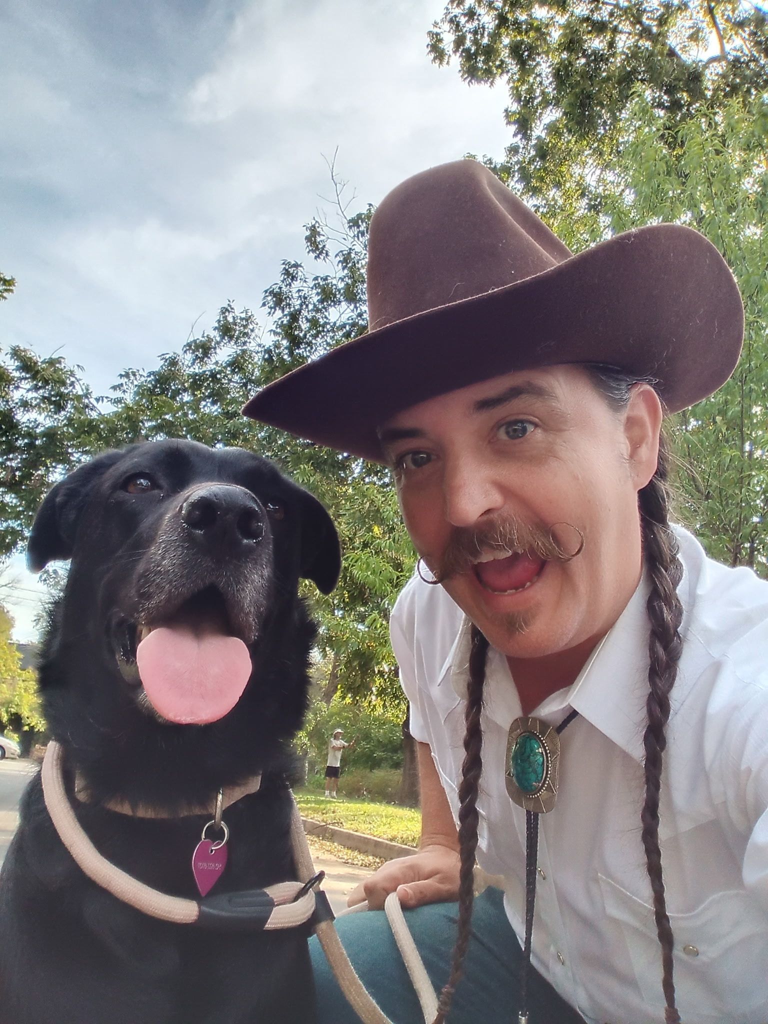 Person in a cowboy hat posing with a black dog outdoors under trees, both facing the camera.