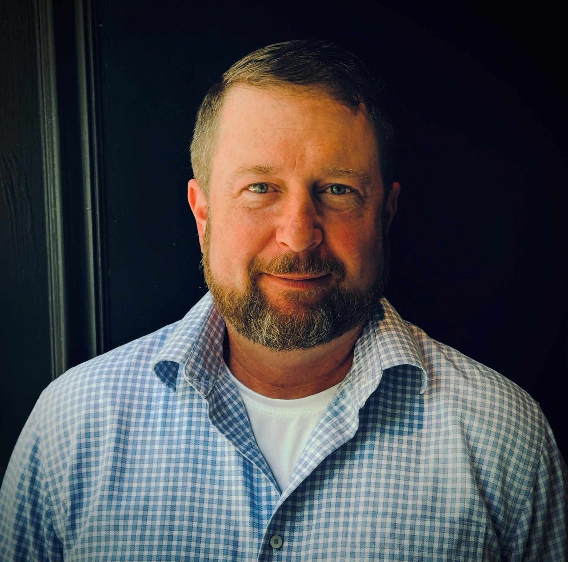 Man with beard smiles, wearing a blue checked shirt over a white undershirt. Against a dark background.