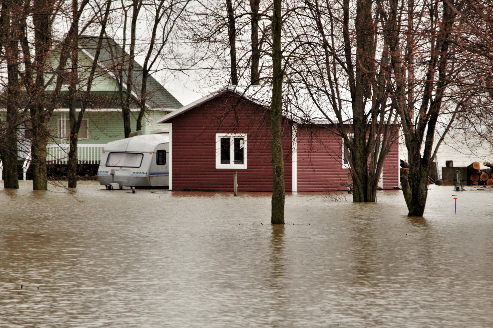 Flooded houses and caravan. Brown water surrounds structures, trees visible in background. Overcast day.