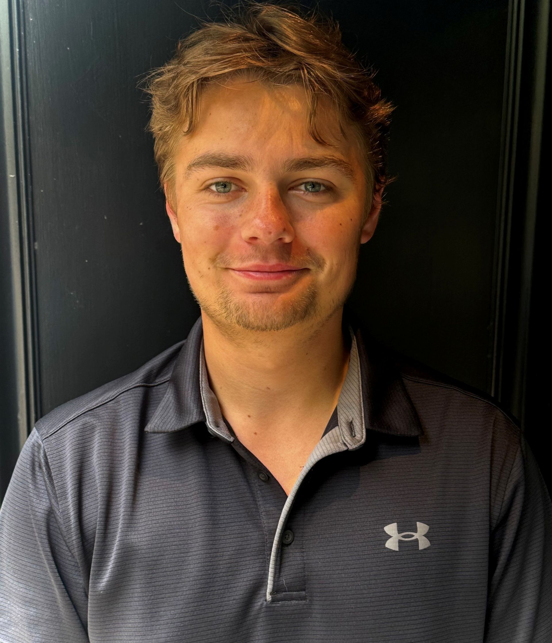 Man with light skin, wearing a dark gray polo shirt, smiles in front of a black wall.