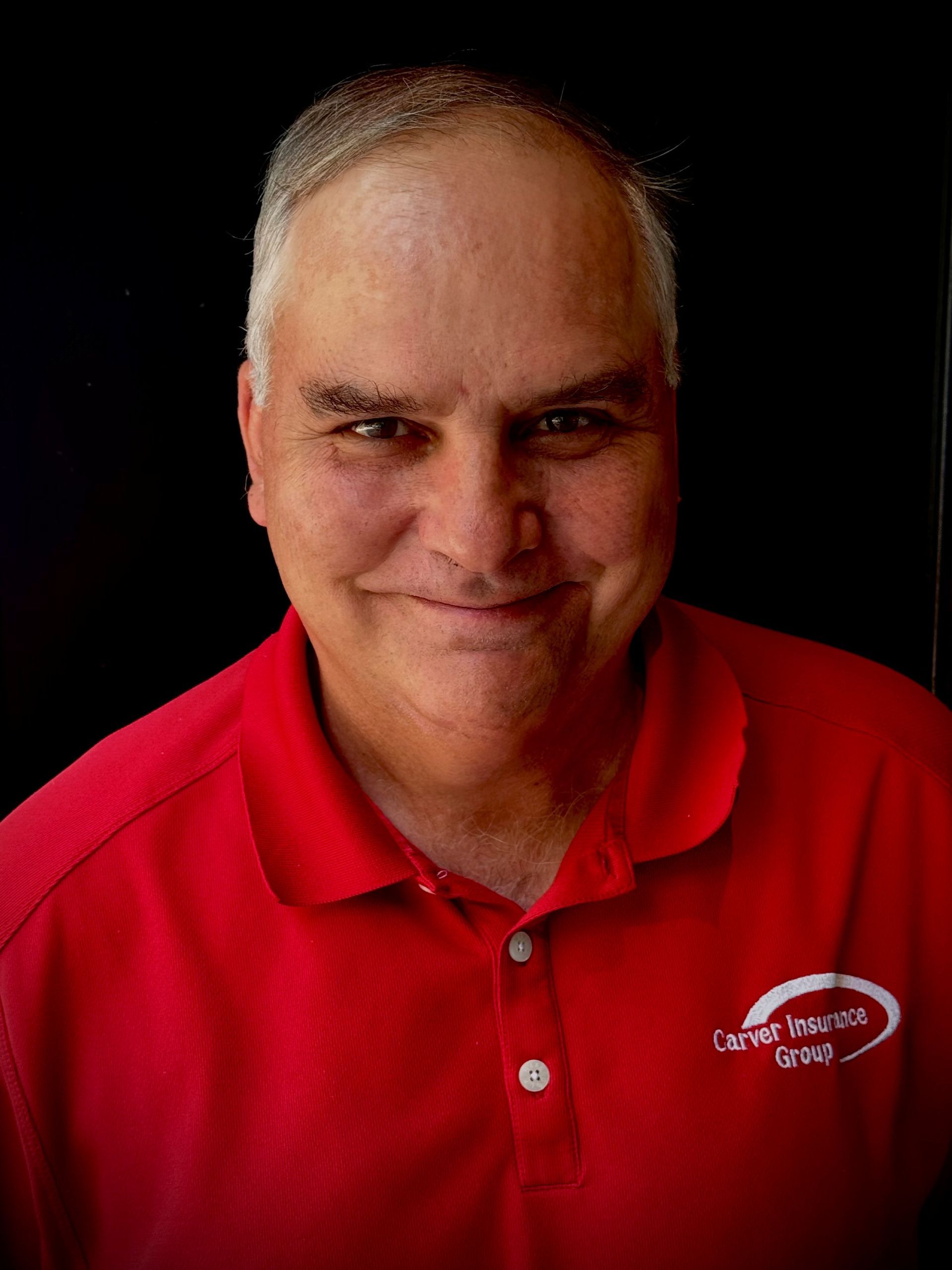 Smiling man in red polo shirt, logo on chest, dark background.