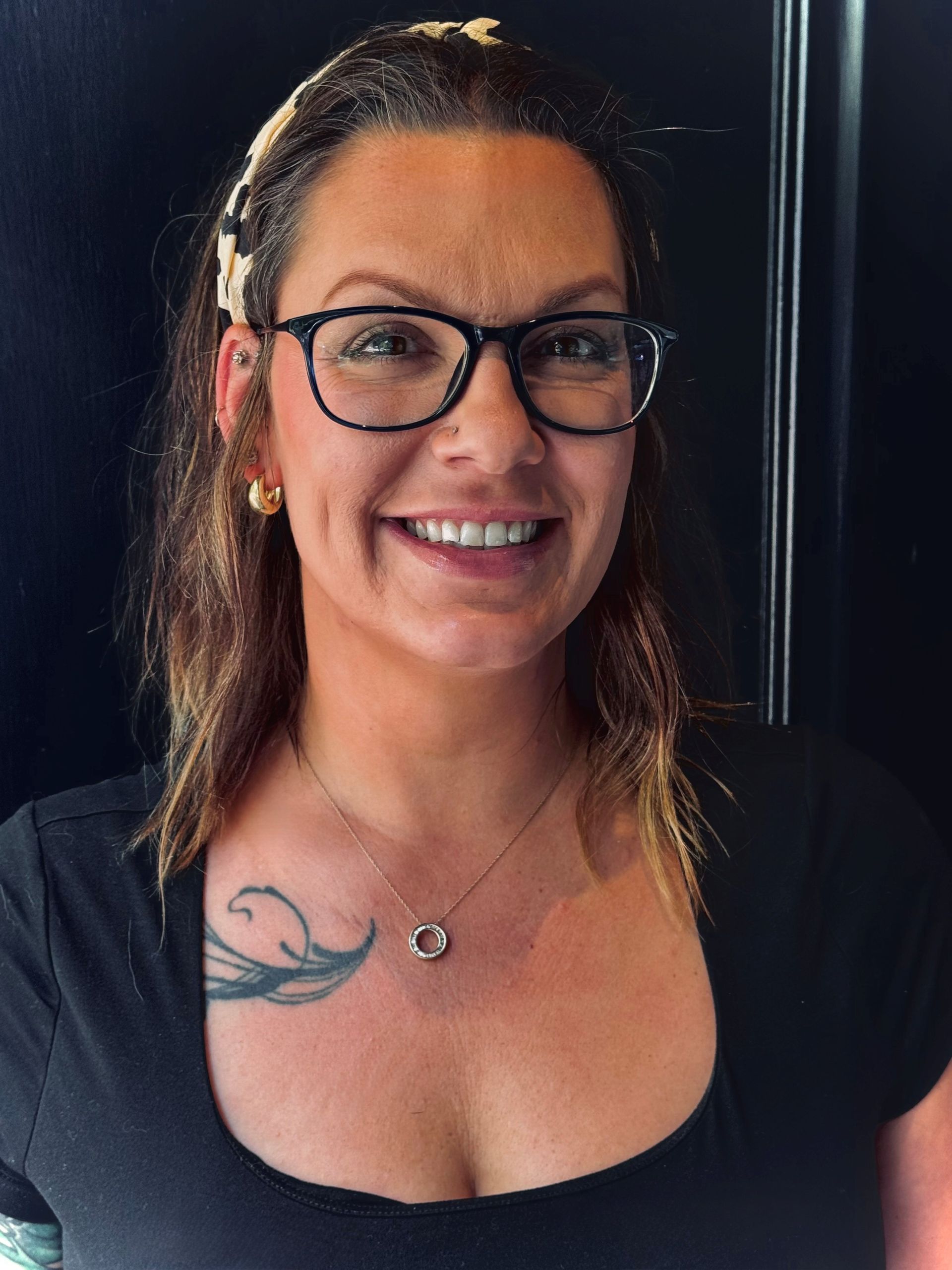 Woman with glasses smiles; tattoo on shoulder; wearing a black shirt, necklace and headband.