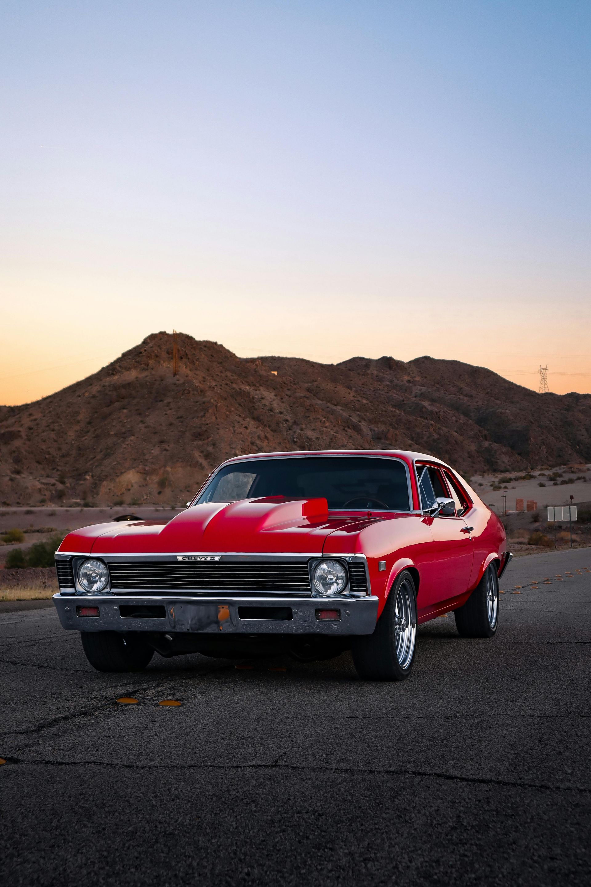 Red classic car parked on asphalt road with a mountain backdrop at sunset.