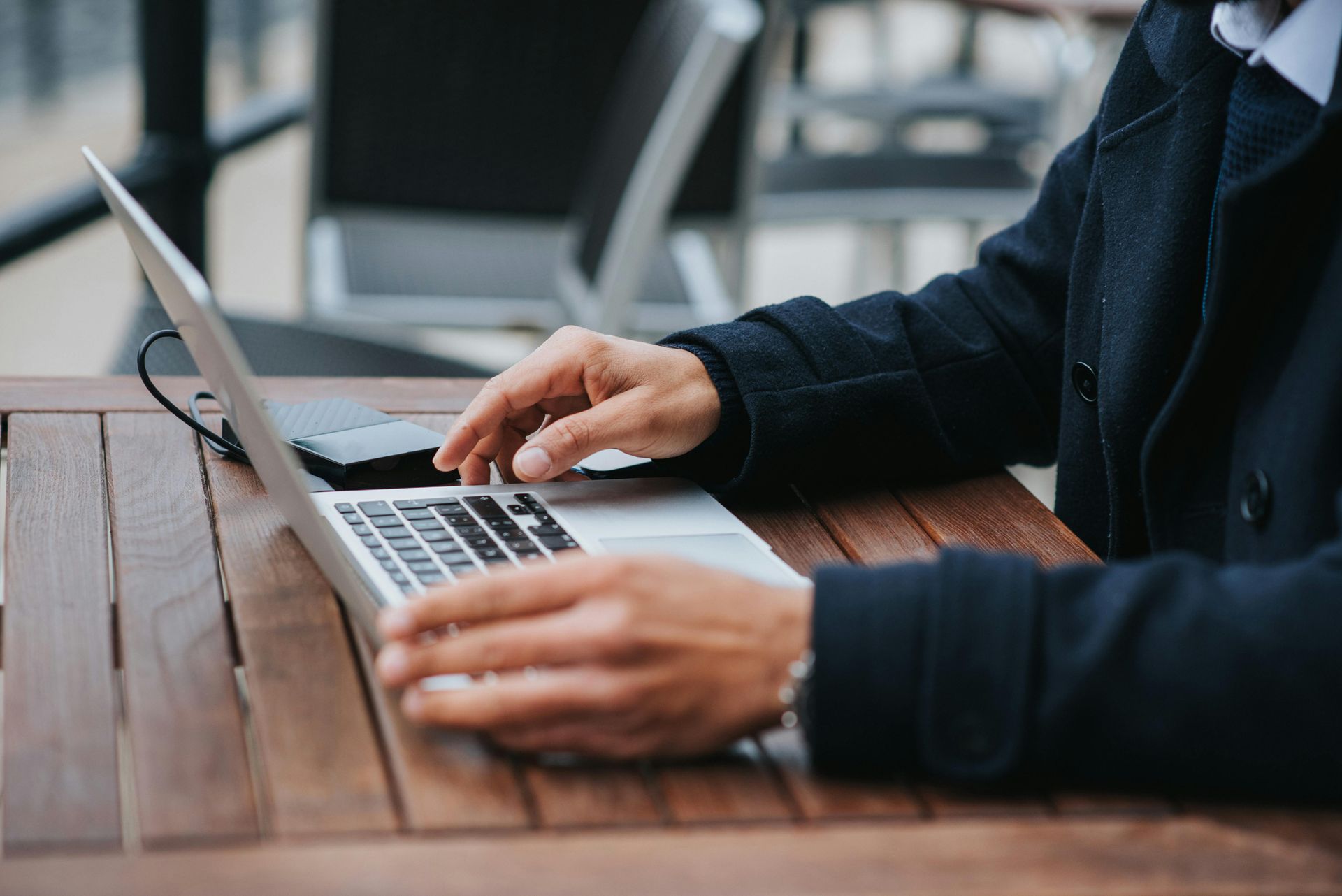Man in a dark coat working on a laptop at a wooden table outdoors.
