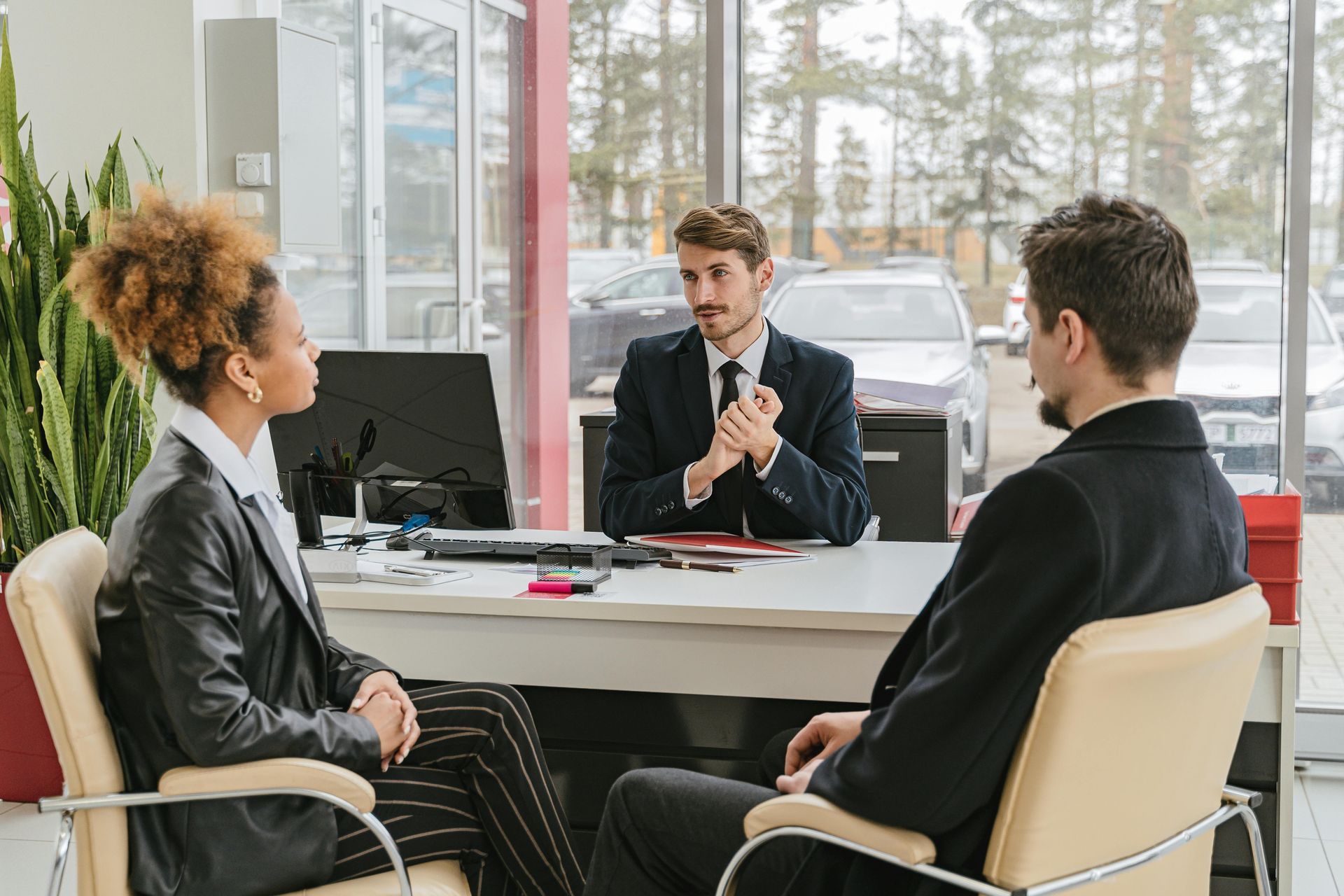 A salesperson with clients in a car dealership, seated at a desk. They are discussing and reviewing documents.