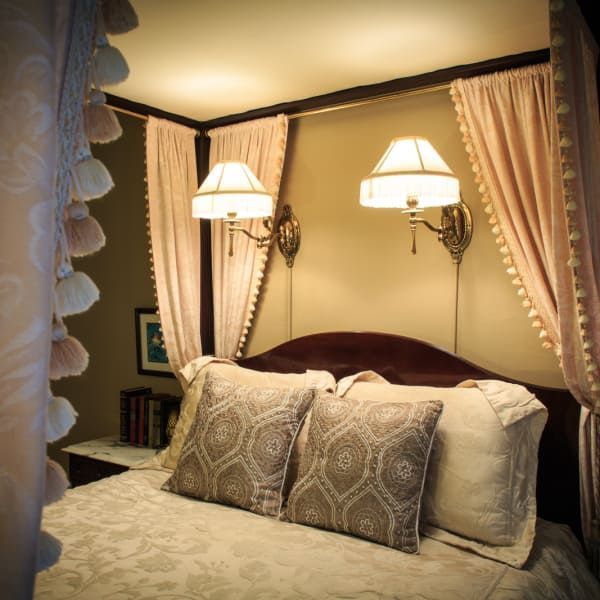Bedroom with canopy bed, pink curtains, sconce lights, patterned pillows, and a nightstand with books.