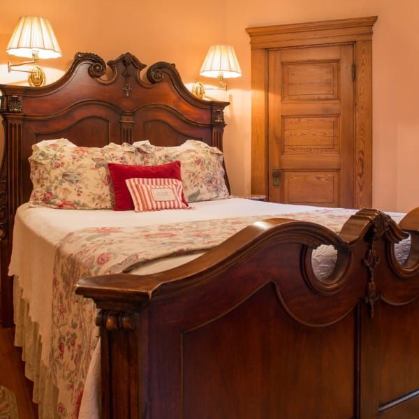 Ornate wooden bed in a bedroom, floral bedding, wall sconces, closed wooden door, peach-colored walls.