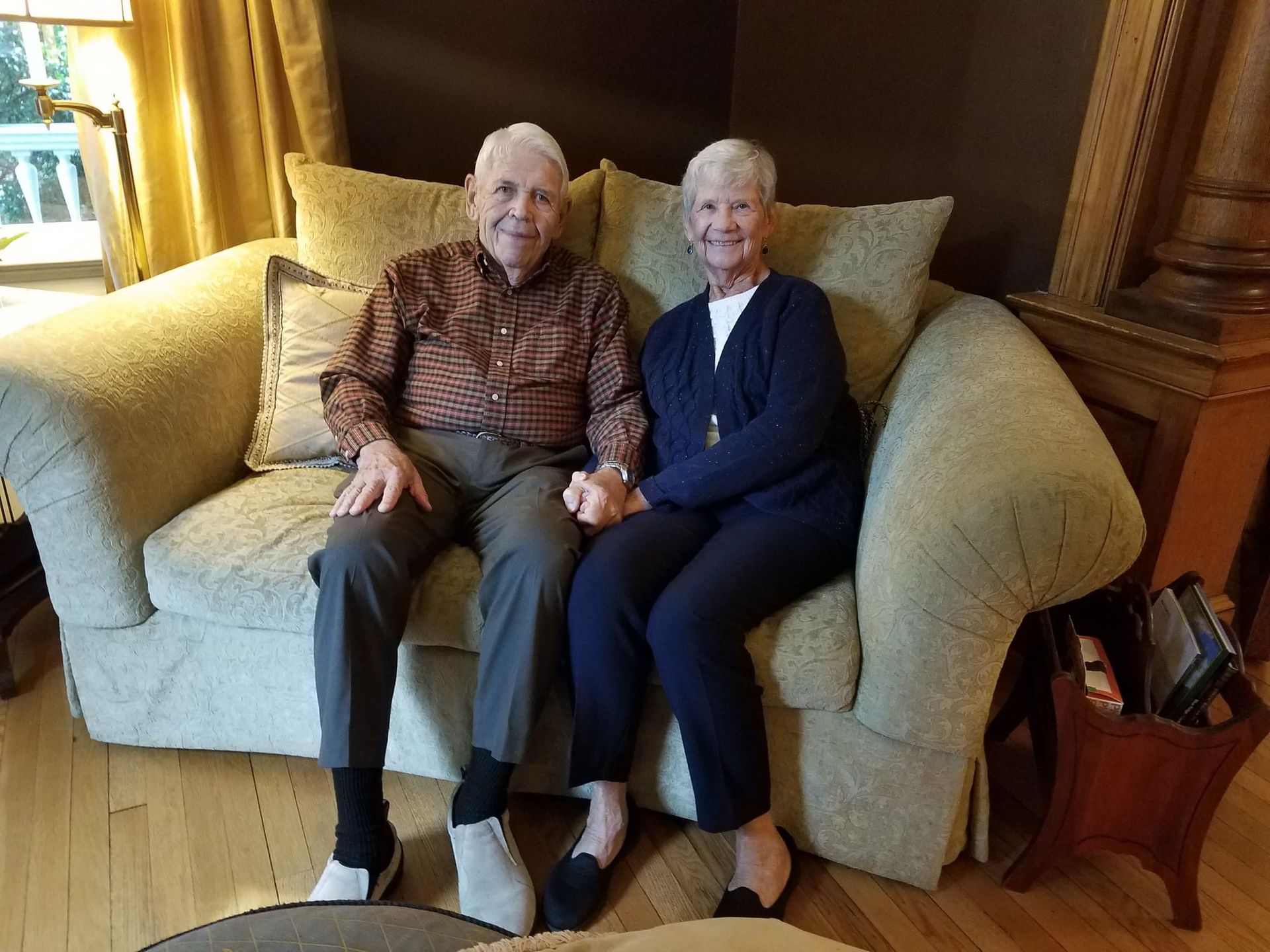 Elderly couple holding hands, sitting on a patterned sofa in a living room, smiling.