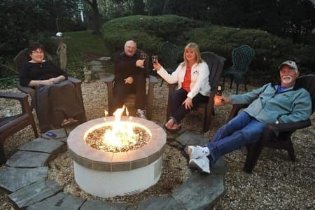 Four people seated around a lit fire pit in a backyard, toasting drinks.