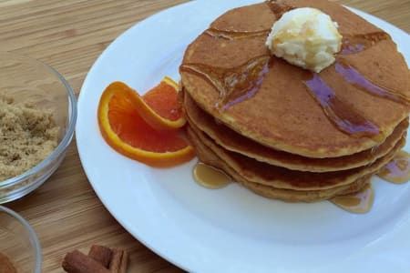 Stack of pancakes with syrup and butter on a white plate, orange slices, and brown sugar.