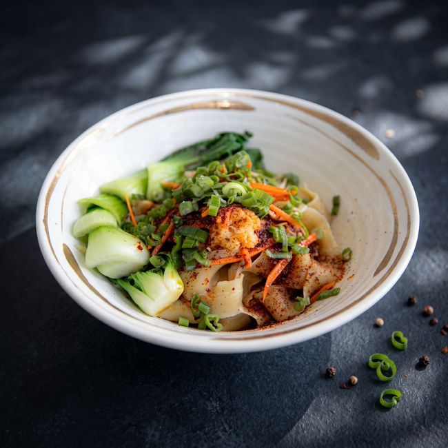 Bowl of noodle soup with greens, chili oil, and herbs on a dark tabletop