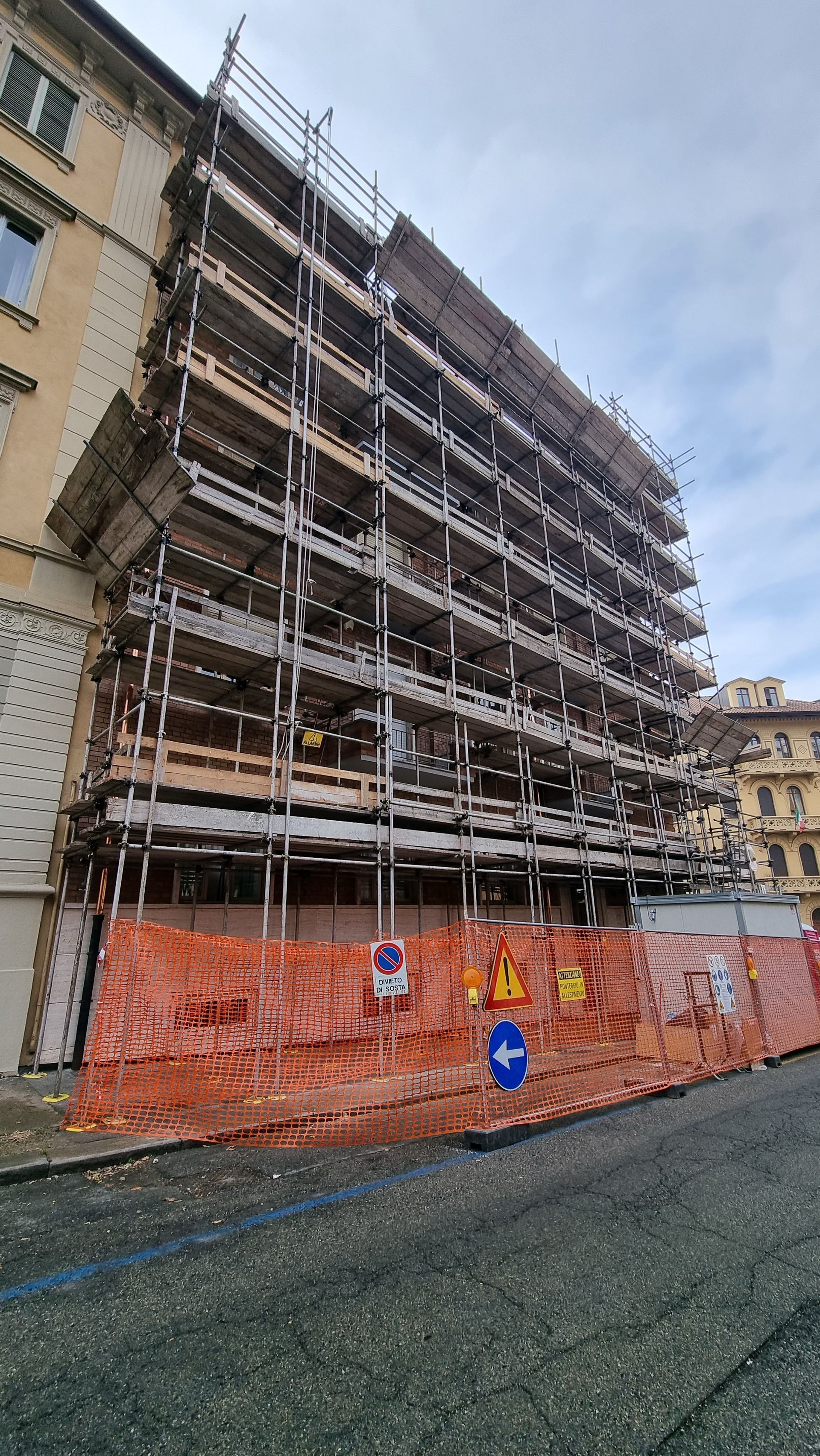 Edificio in fase di ristrutturazione, ricoperto da impalcature, con reti di sicurezza arancioni lungo la strada.