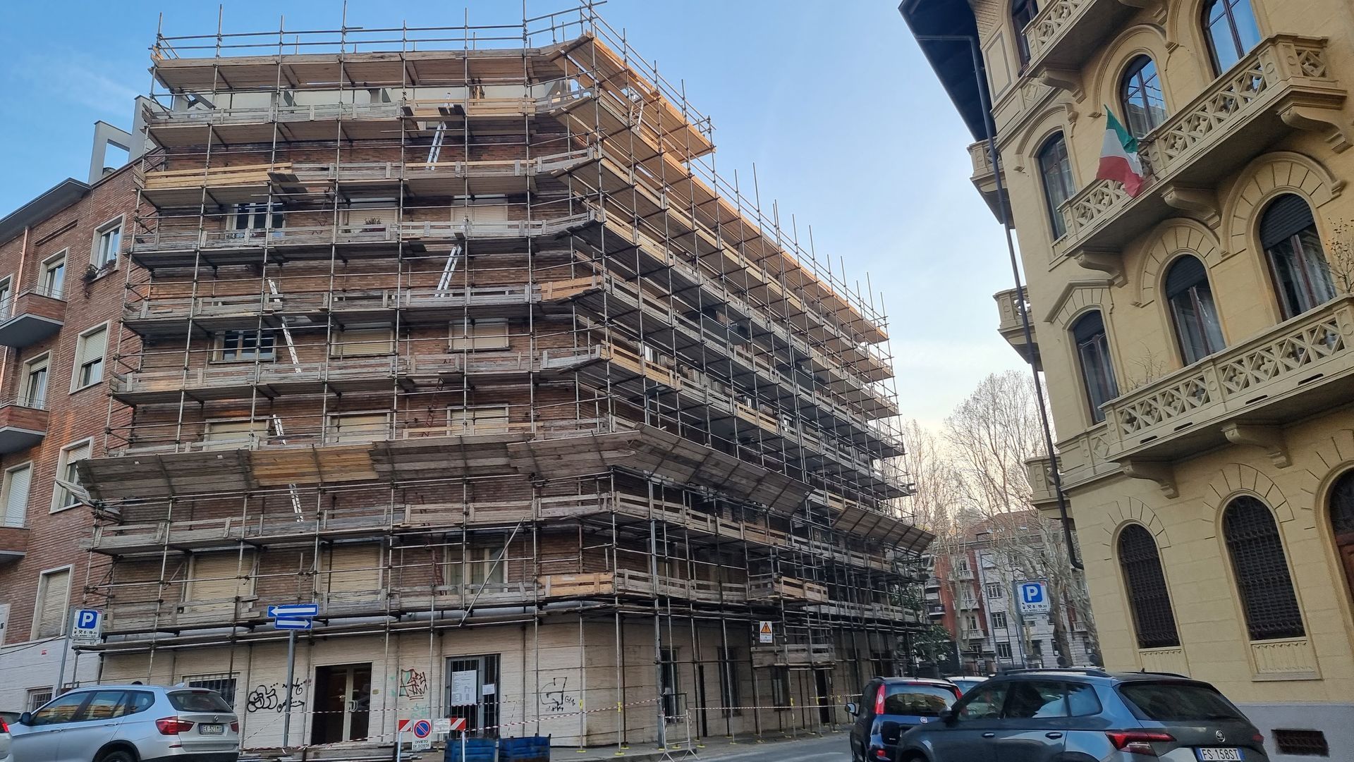 Edificio d'angolo ricoperto di impalcature su una via cittadina, con auto parcheggiate e un palazzo di appartamenti beige accanto.