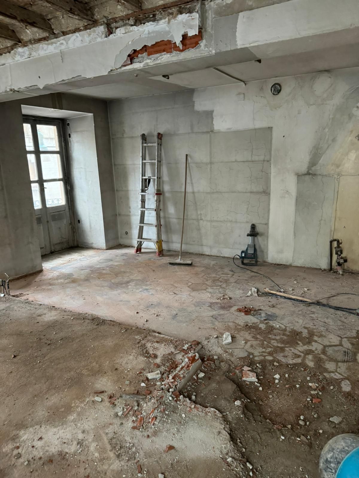 Stanza vuota e non rifinita con intonaco scrostato, detriti sul pavimento, una scala appoggiata al muro e una porta sulla sinistra.