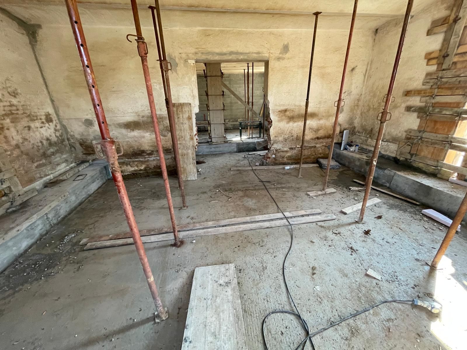 Stanza ristrutturata con impalcature, pareti non rifinite e cavi elettrici a vista su un pavimento in cemento.