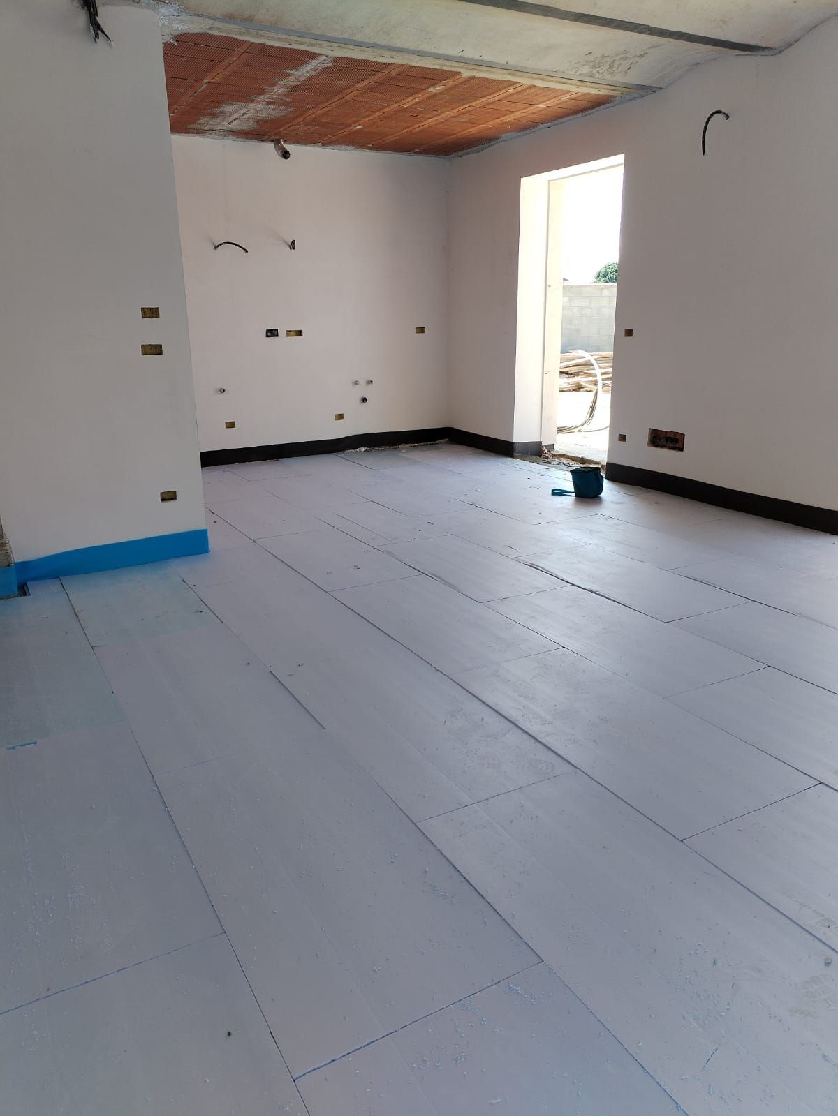 Stanza vuota e non rifinita con pareti bianche, soffitto a vista e porta aperta che lascia entrare una luce intensa.