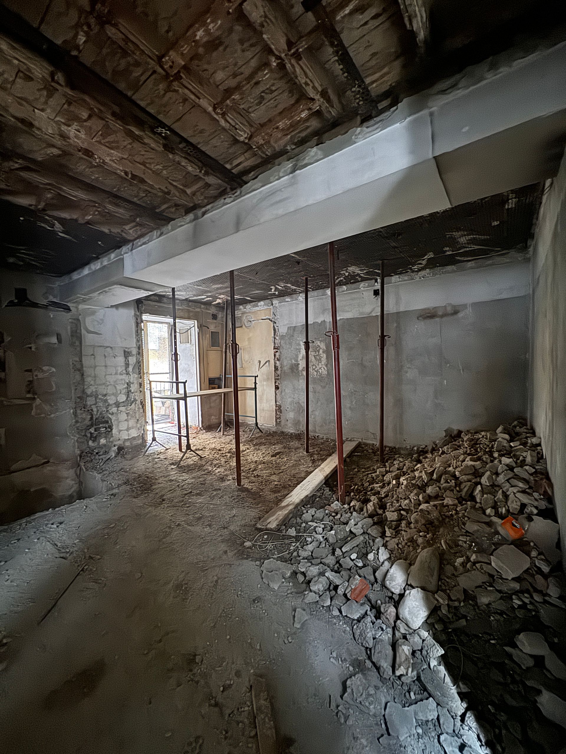 Stanza parzialmente demolita con cumuli di macerie, travi a vista e una porta che dà su un esterno luminoso.