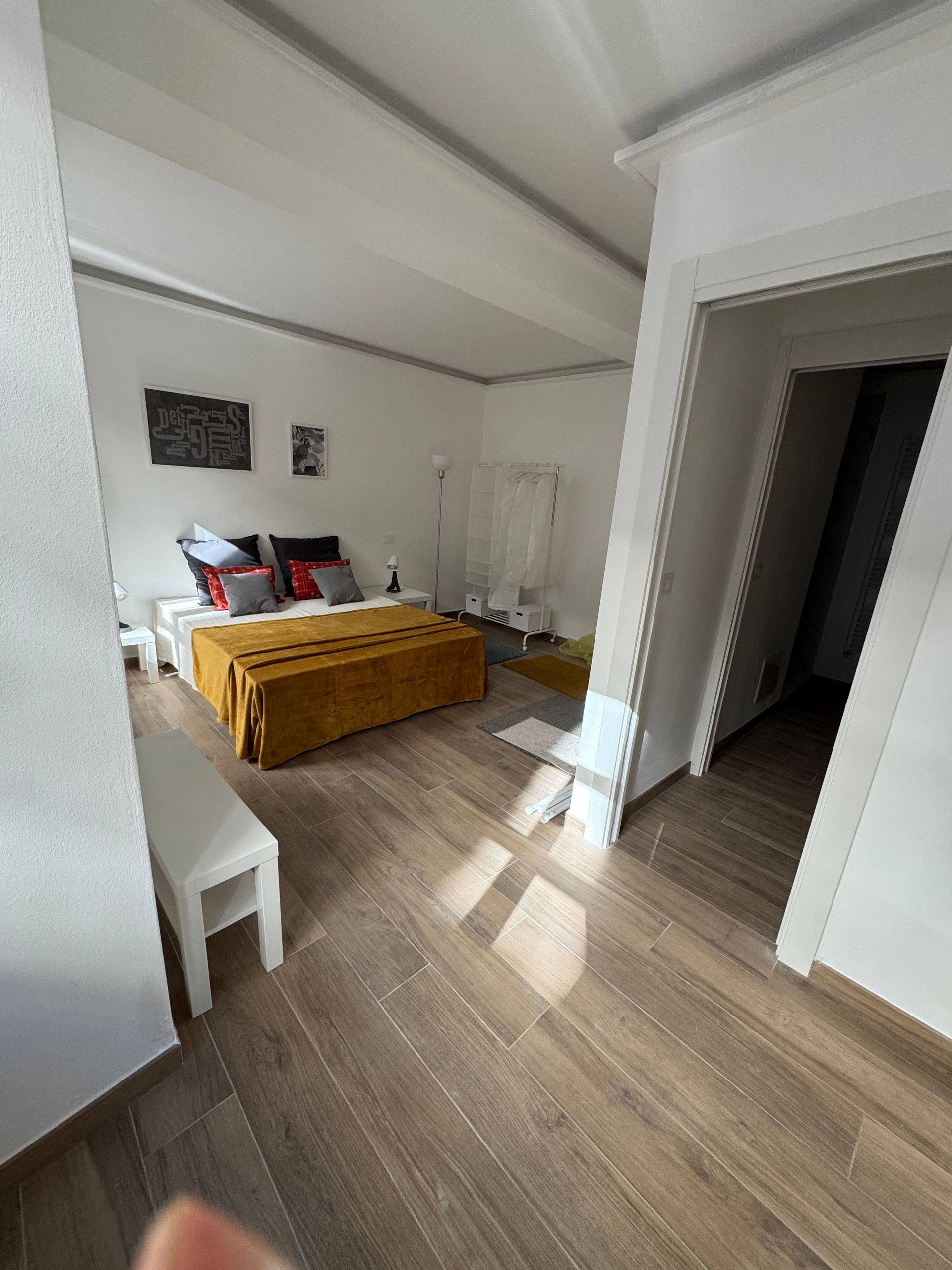 Camera da letto luminosa con letto, coperta color senape, sedia bianca e luce del sole che filtra sul pavimento in legno.