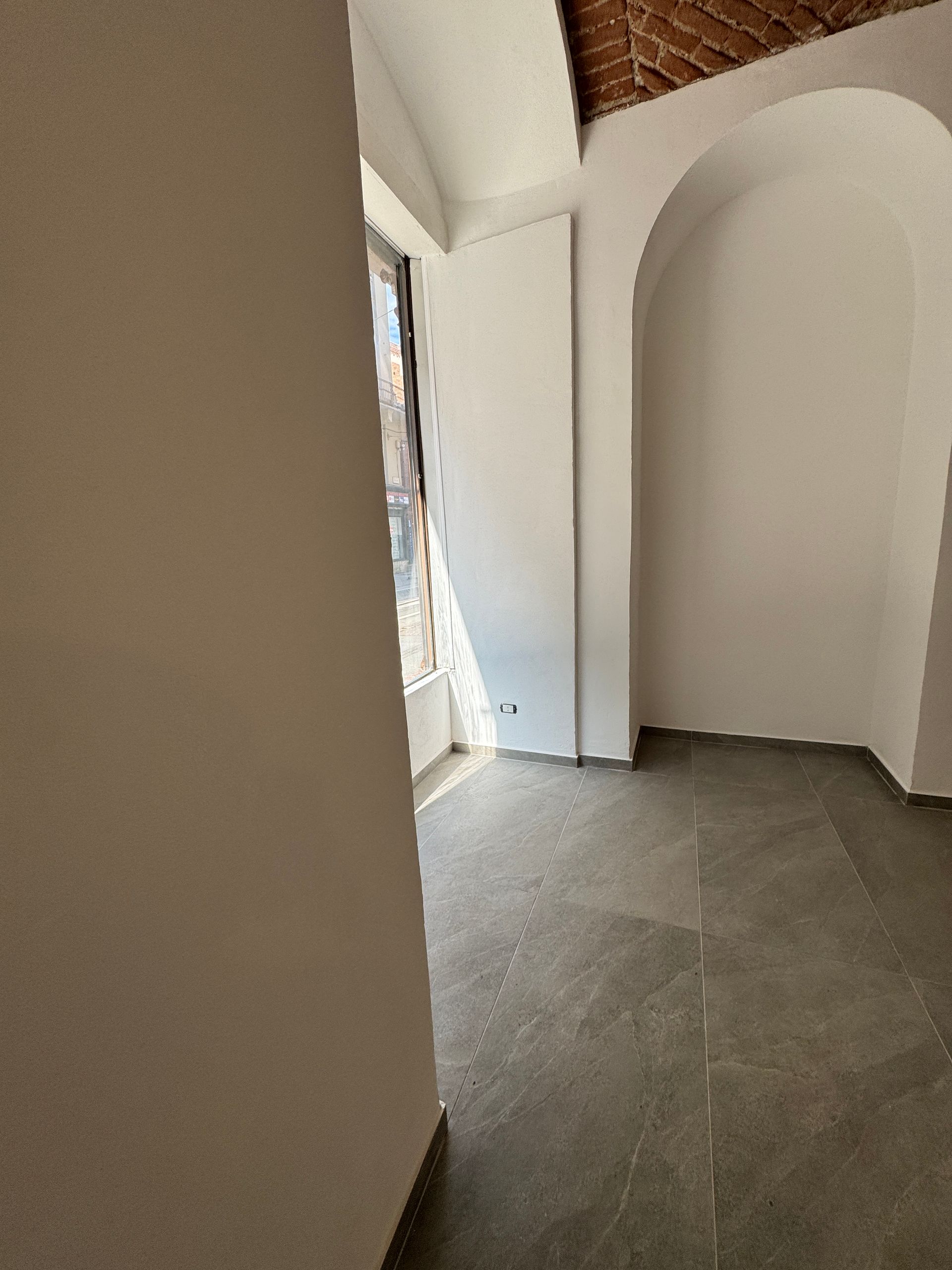 Corridoio curvo bianco con nicchia ad arco, pavimento in piastrelle grigie e una porta che si apre sulla luce naturale.