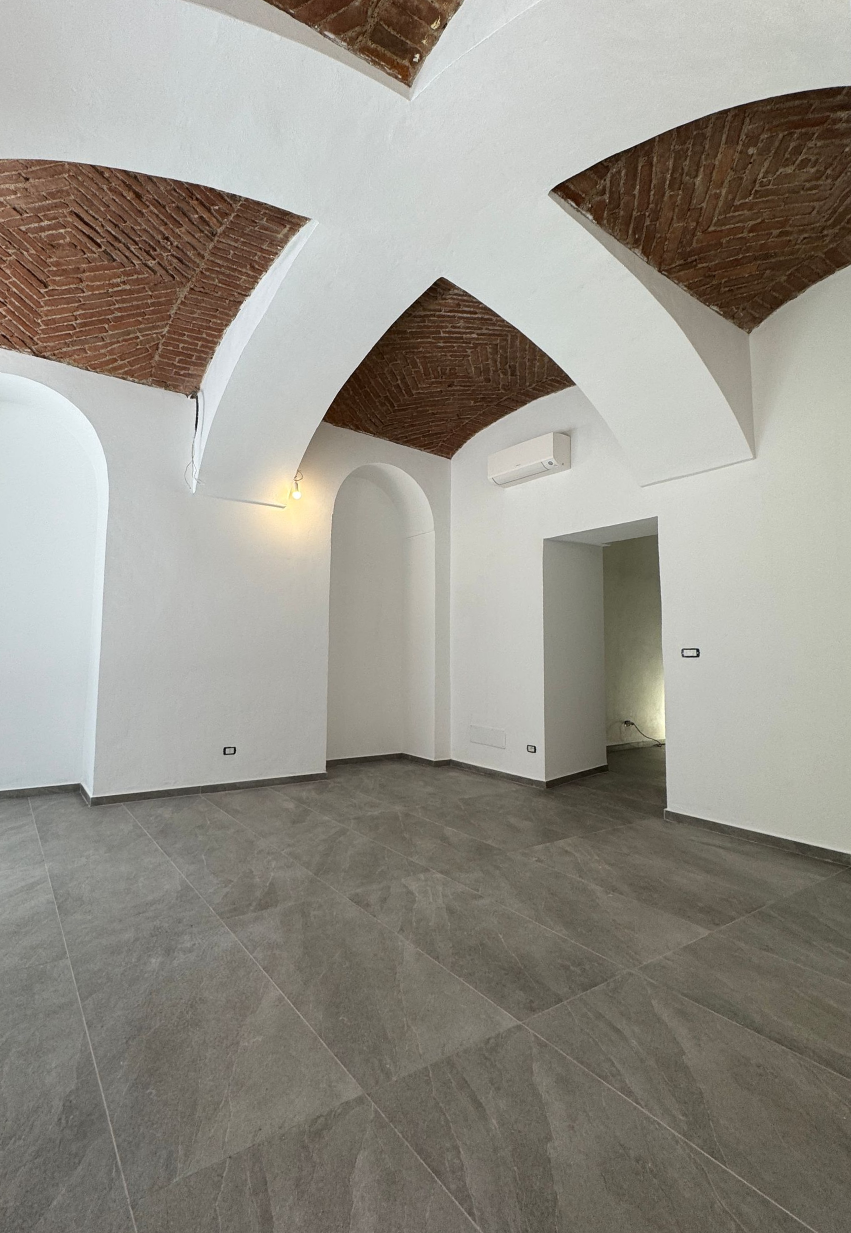 Stanza bianca vuota con soffitto a volta in mattoni e pavimento in piastrelle grigie.