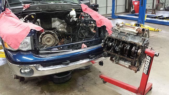 Blue truck with exposed engine bay, next to a dissembled engine on a stand in a garage.