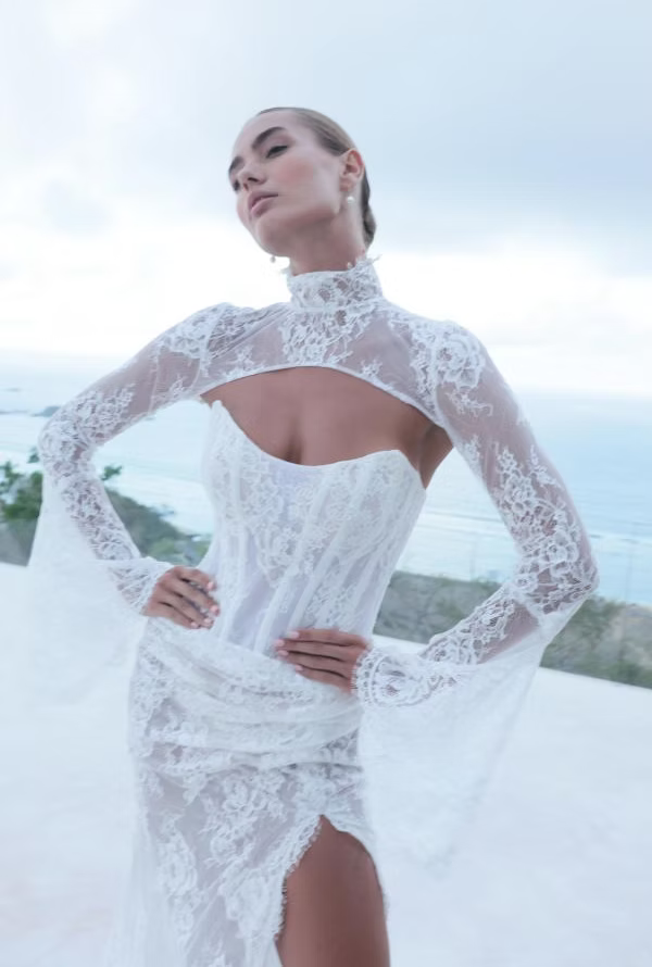 Woman in white lace wedding dress with high neck, arms akimbo, outdoors.