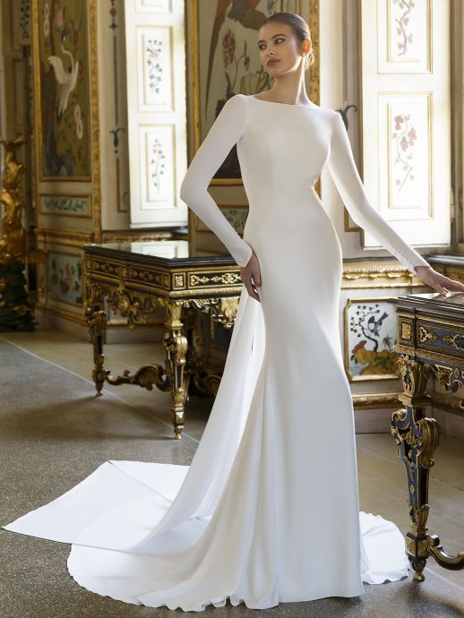 Woman in a white, long-sleeved wedding dress with a train, standing in an ornate room.