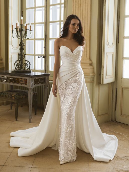 Woman in a strapless white wedding dress with a train, standing in a room with large windows.
