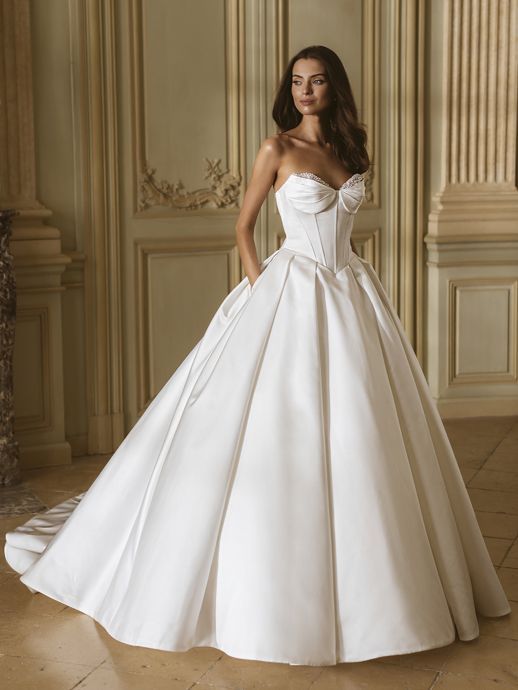 Woman in a white strapless ballgown with train, standing indoors, hands in pockets.