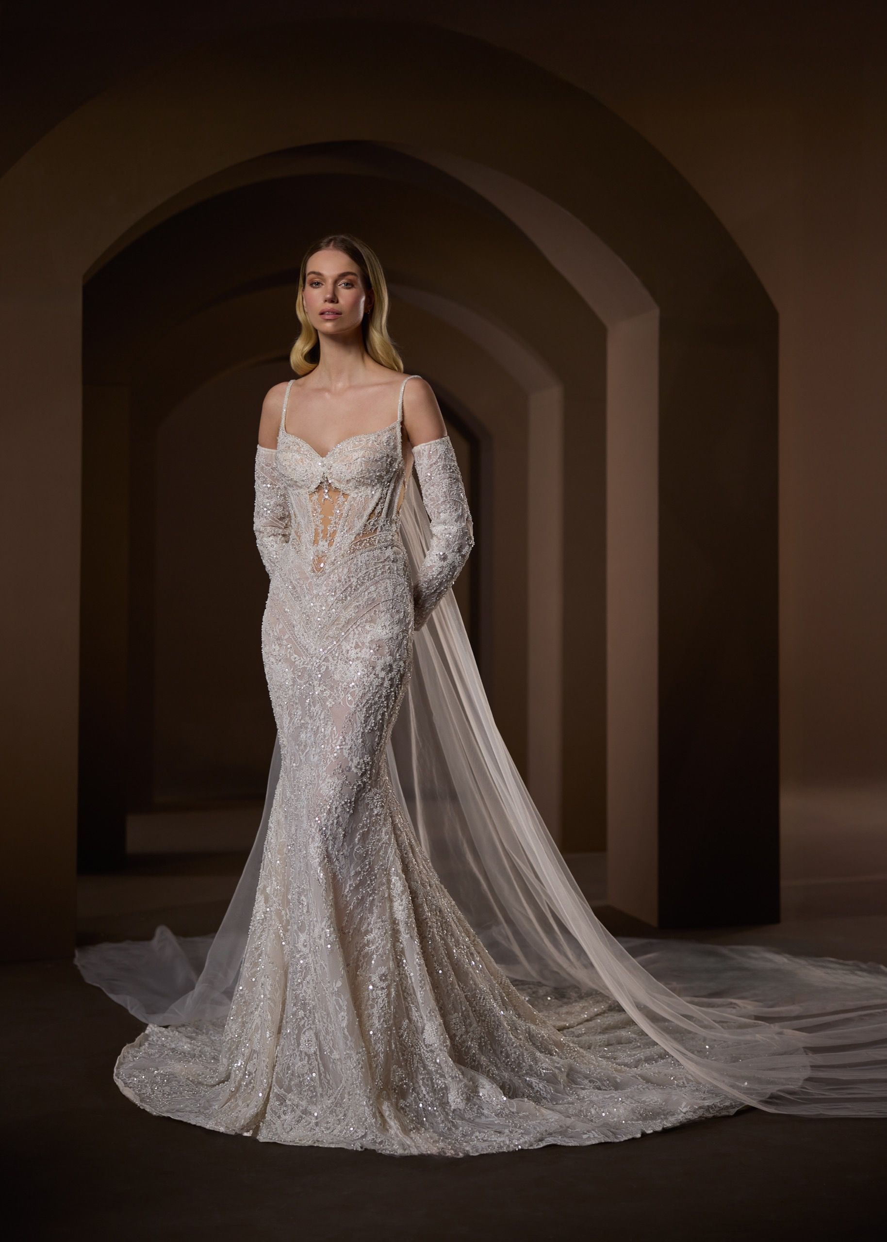 Woman in a sparkling white wedding gown with a long train and sheer sleeves, in a dimly lit hallway.
