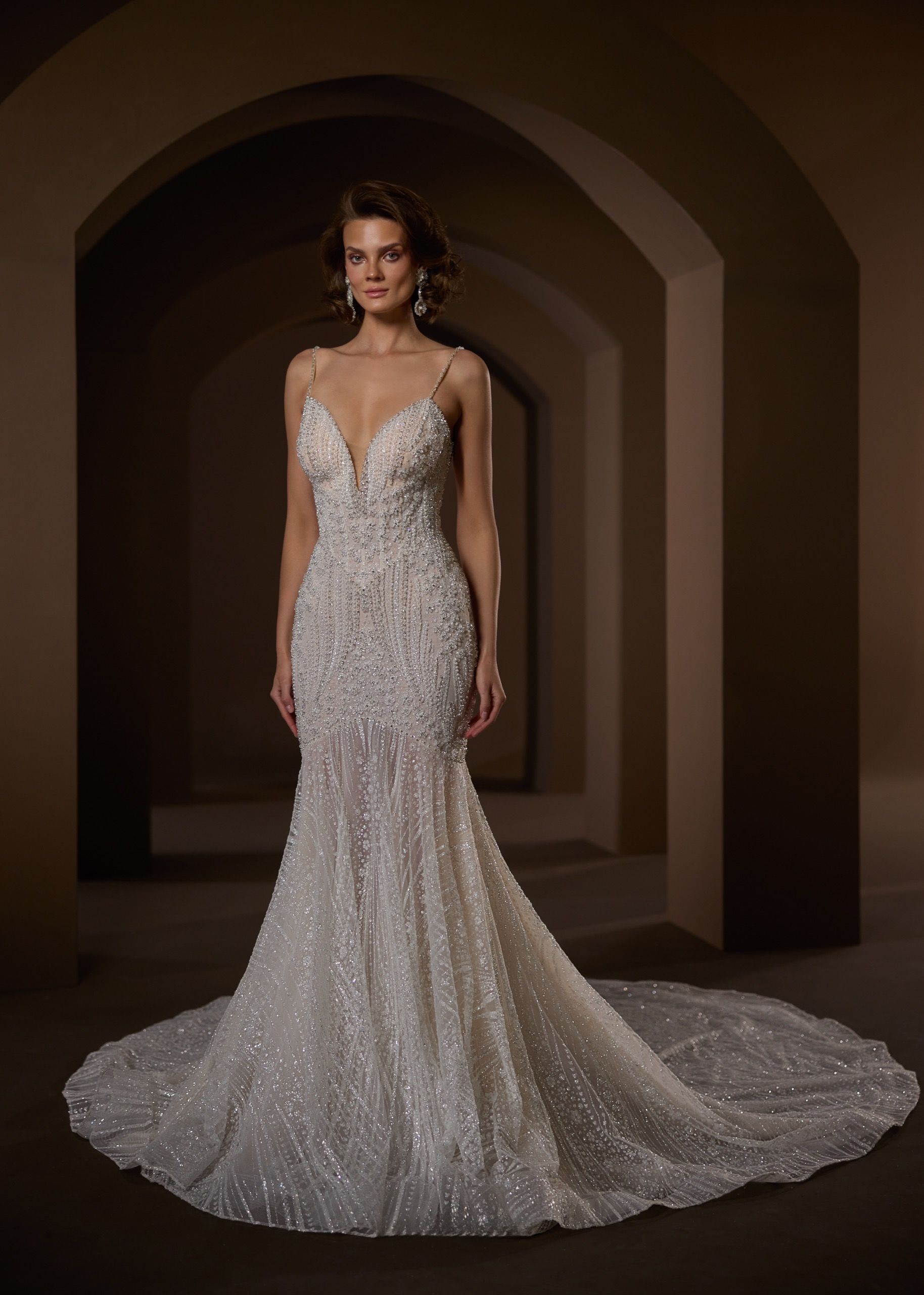 Woman in a beaded, fitted wedding dress with a train, standing in a dark arched hallway.