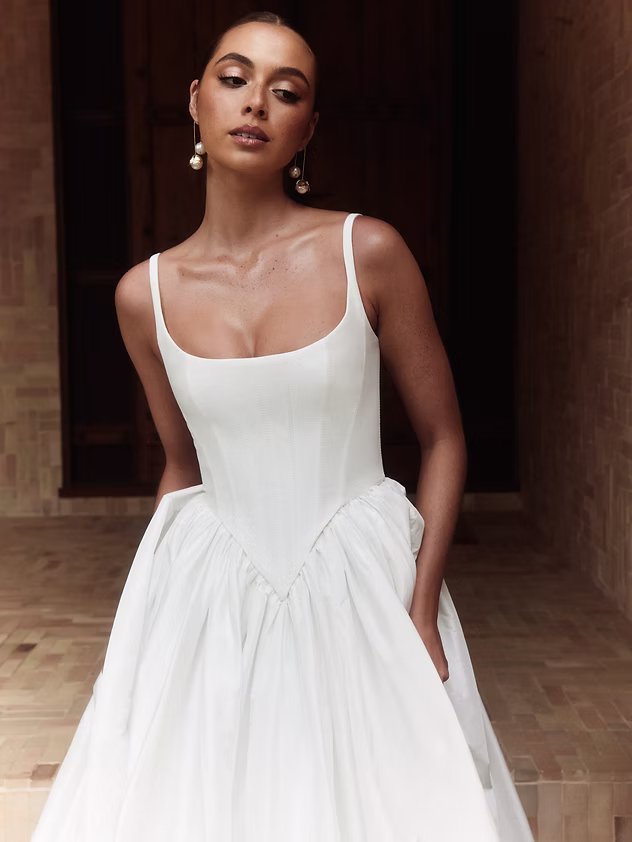 Woman in white bridal gown, pearls, square neckline, standing, looking forward.
