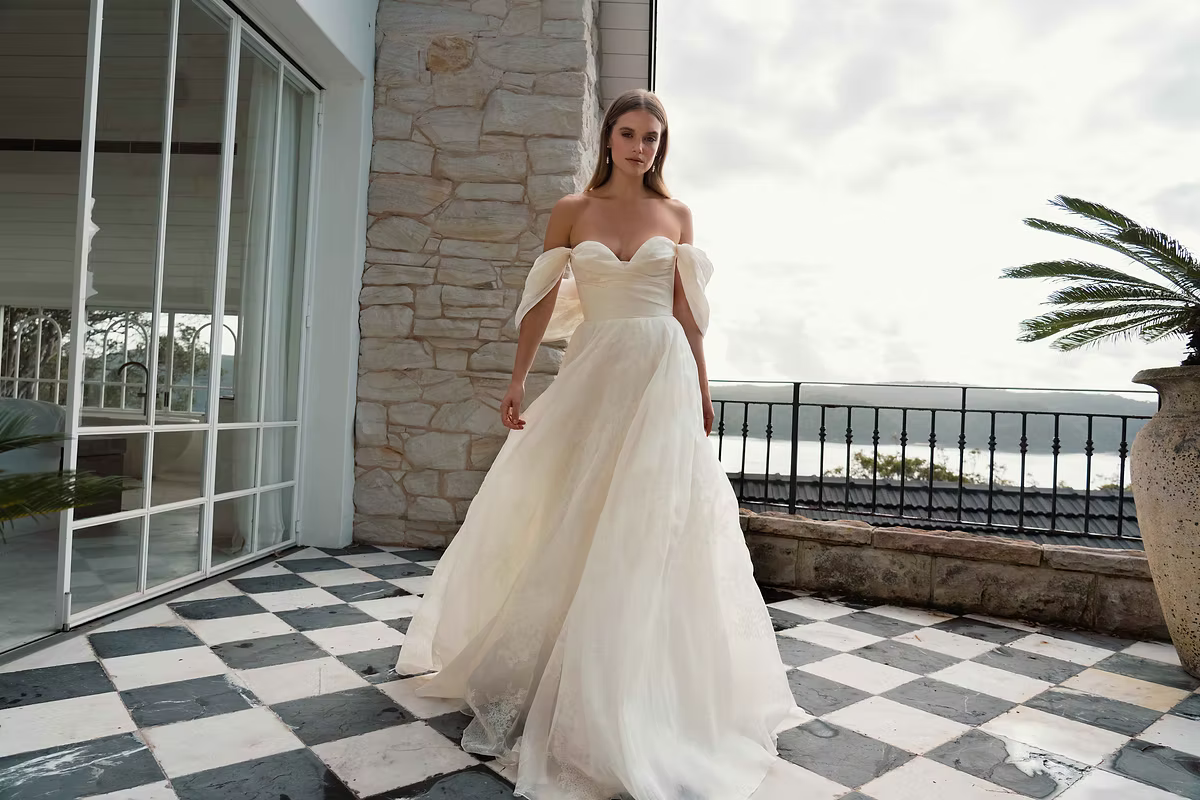 Woman in off-shoulder wedding dress, standing on a checkered patio overlooking water.