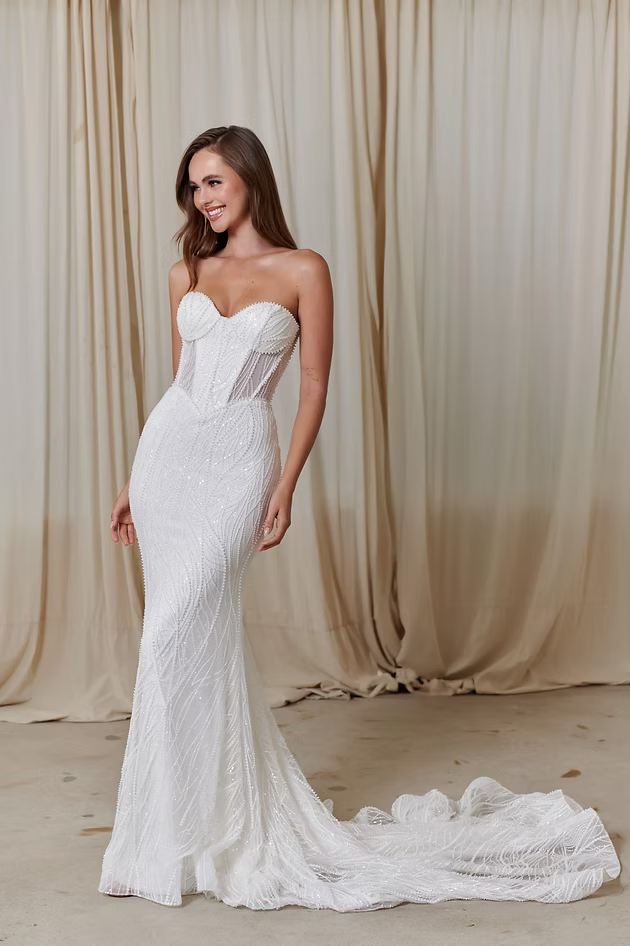 Woman in white strapless wedding dress, smiling, with long train, standing in front of curtains.