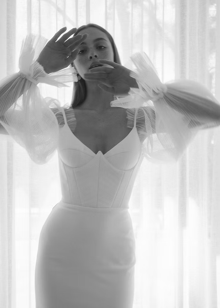 Woman in white wedding dress, sheer puff sleeves, hands near face.
