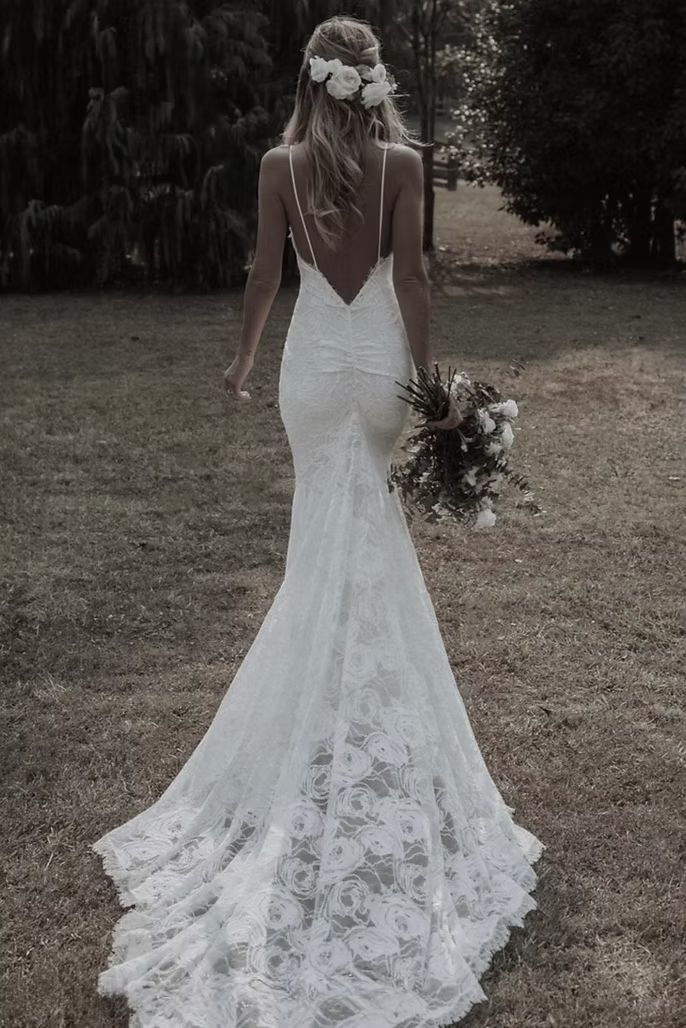 Bride in a lace wedding dress with a low back, holding a bouquet, walking outdoors.
