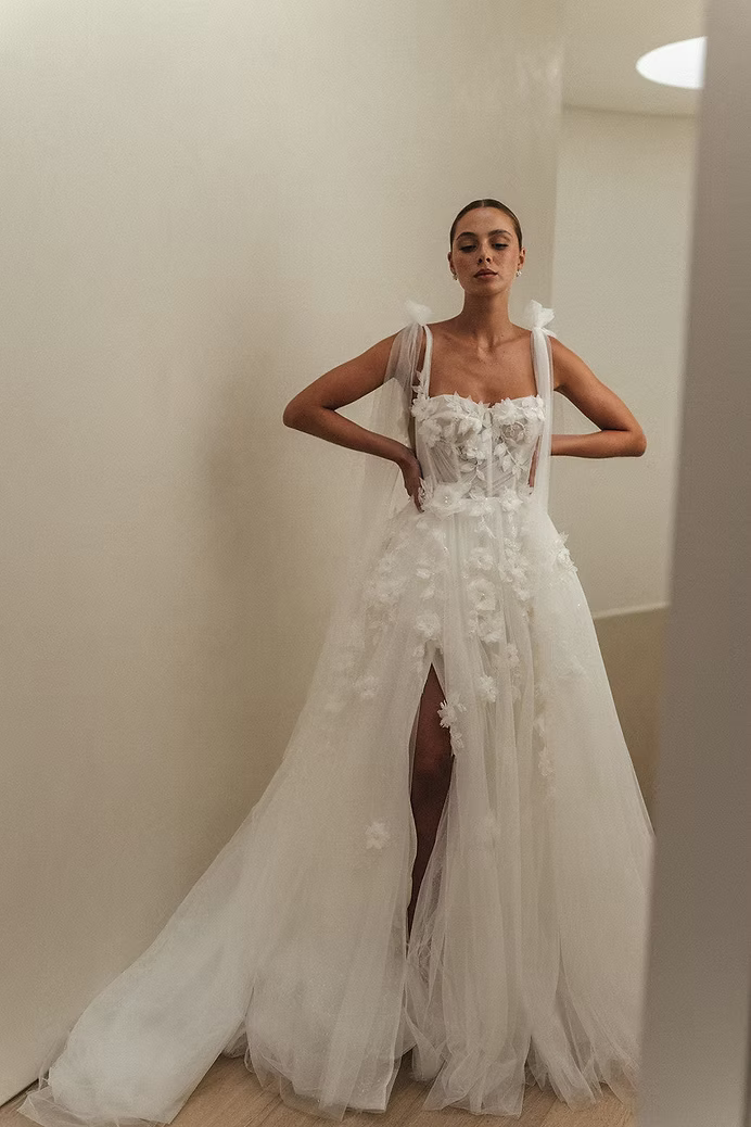 Woman in white floral wedding gown with a high slit, posing indoors.