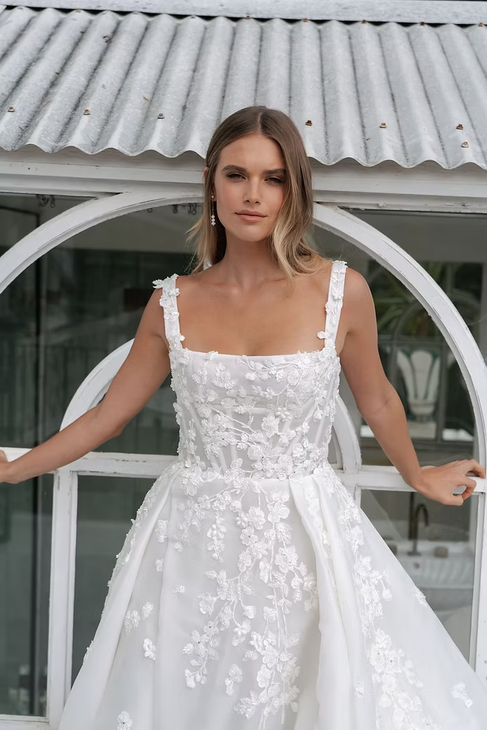 Woman in white wedding dress poses in front of a window, arms outstretched.