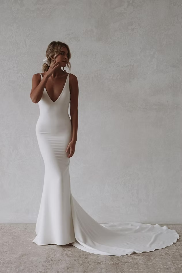 Woman in white wedding dress with low neckline and long train, standing in front of a neutral wall.