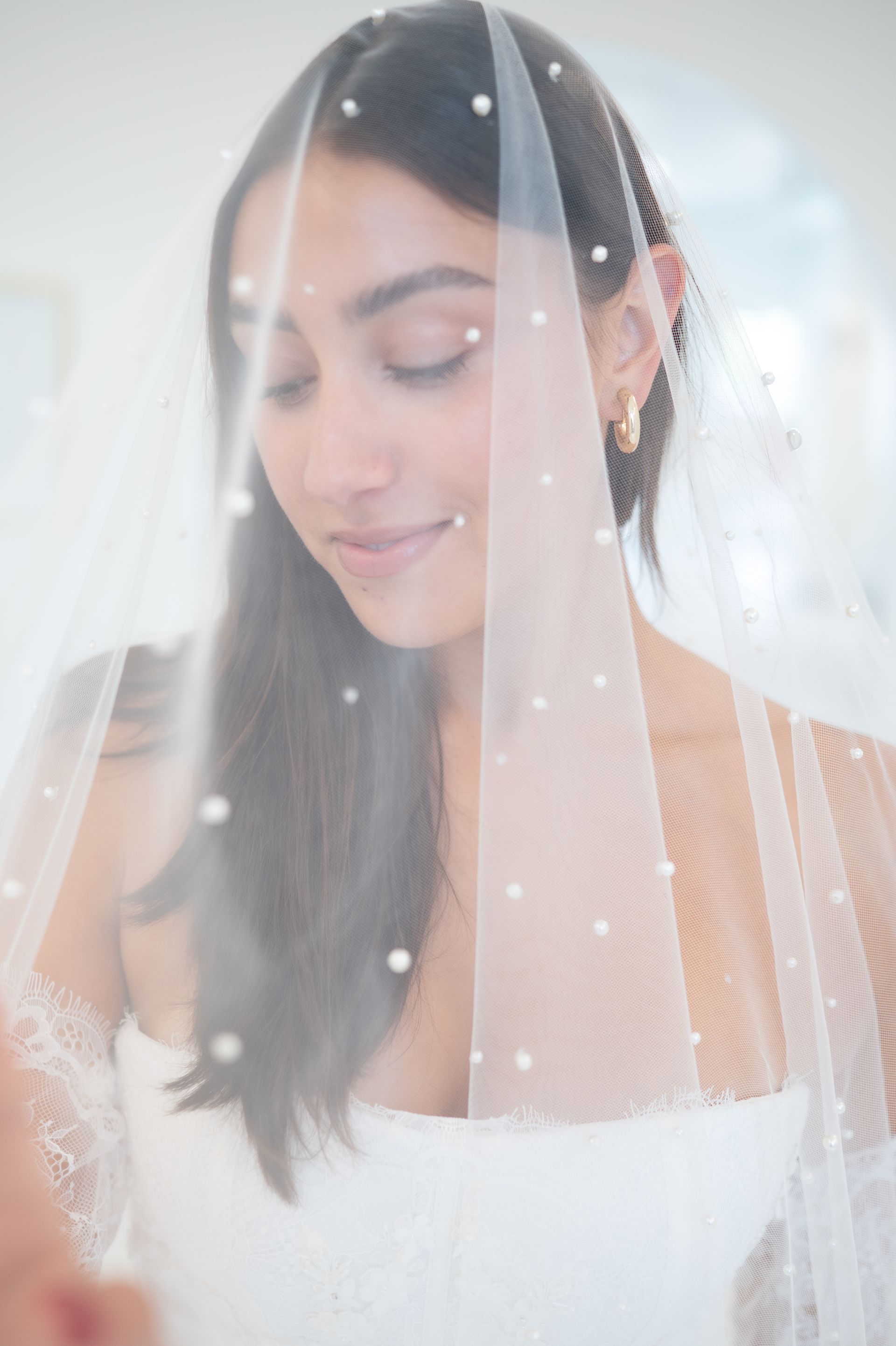 Woman with a pearl-studded veil, smiling with closed eyes, wearing gold hoop earrings.