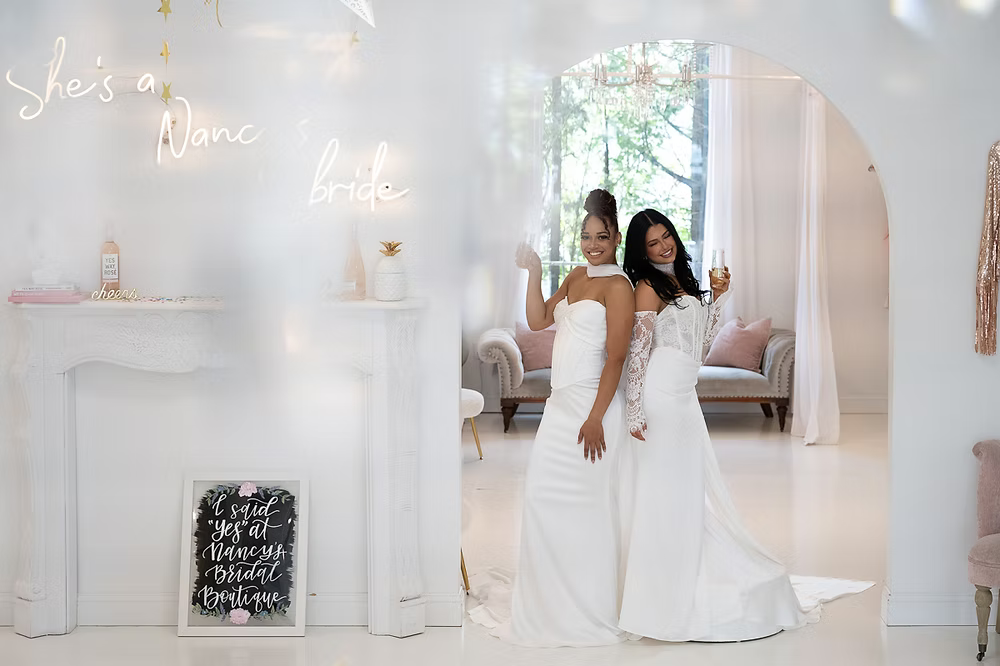 Two women in white wedding dresses, smiling, pose near a fireplace, bright setting, lit archway.