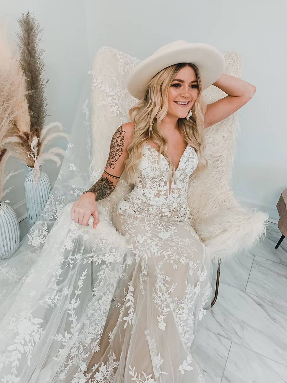 Woman in wedding dress and hat, seated in a chair, arm raised, smiling.